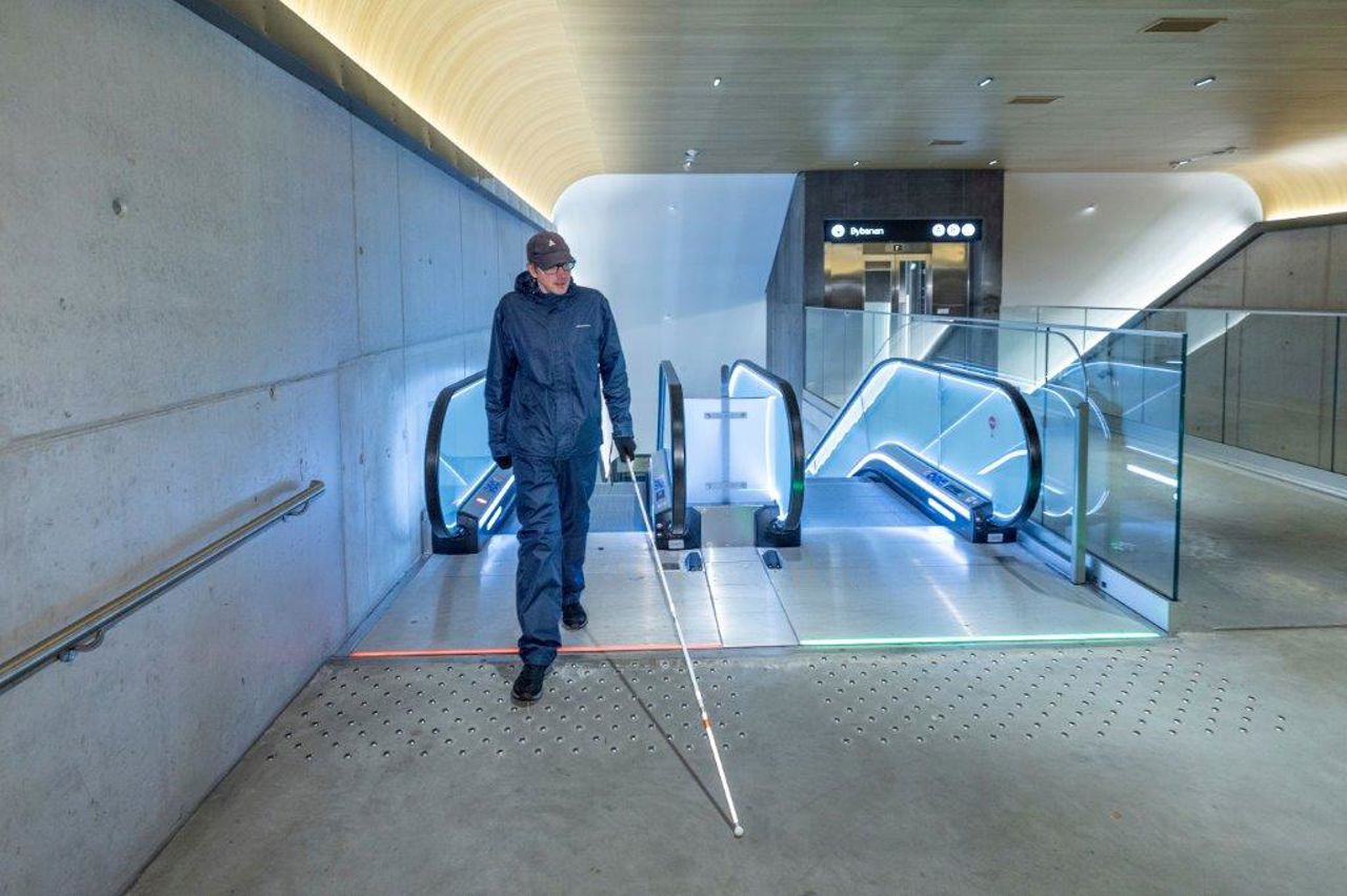 Biletet syner ein mann som kjem opp ein rulletrapp med ei raud lysstripe foran rulletrappen. Rulletrappen ved sisa av har ein grøn strek som viser at denen rulletrappen kan nyttast for å kome seg ned til haldeplassen.