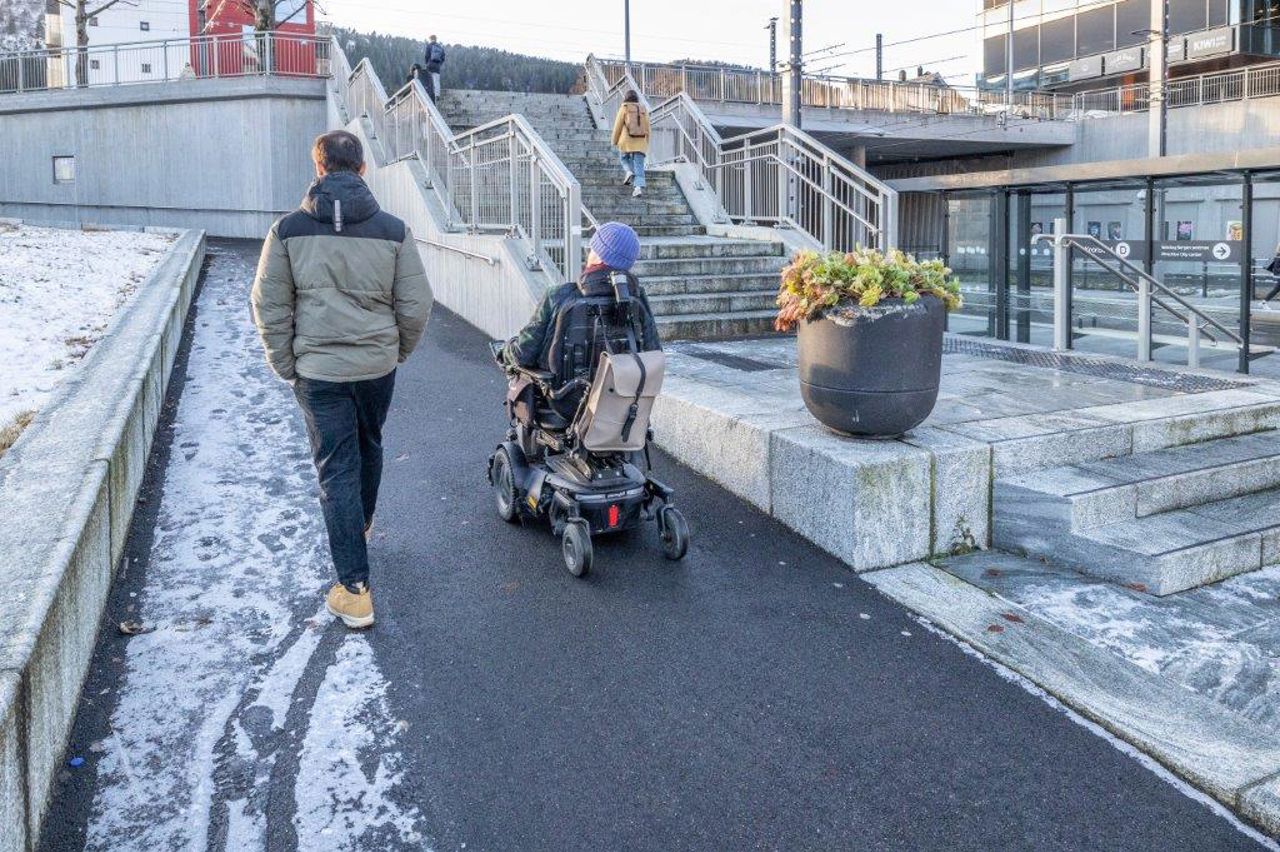 Biletet syner ein mann som køyrer rullestol på bar asfalt vinterstid på grunn av at det er varmekablar i bakken mellom haldeplassane på linje 1 og linje 2 på Kronstad