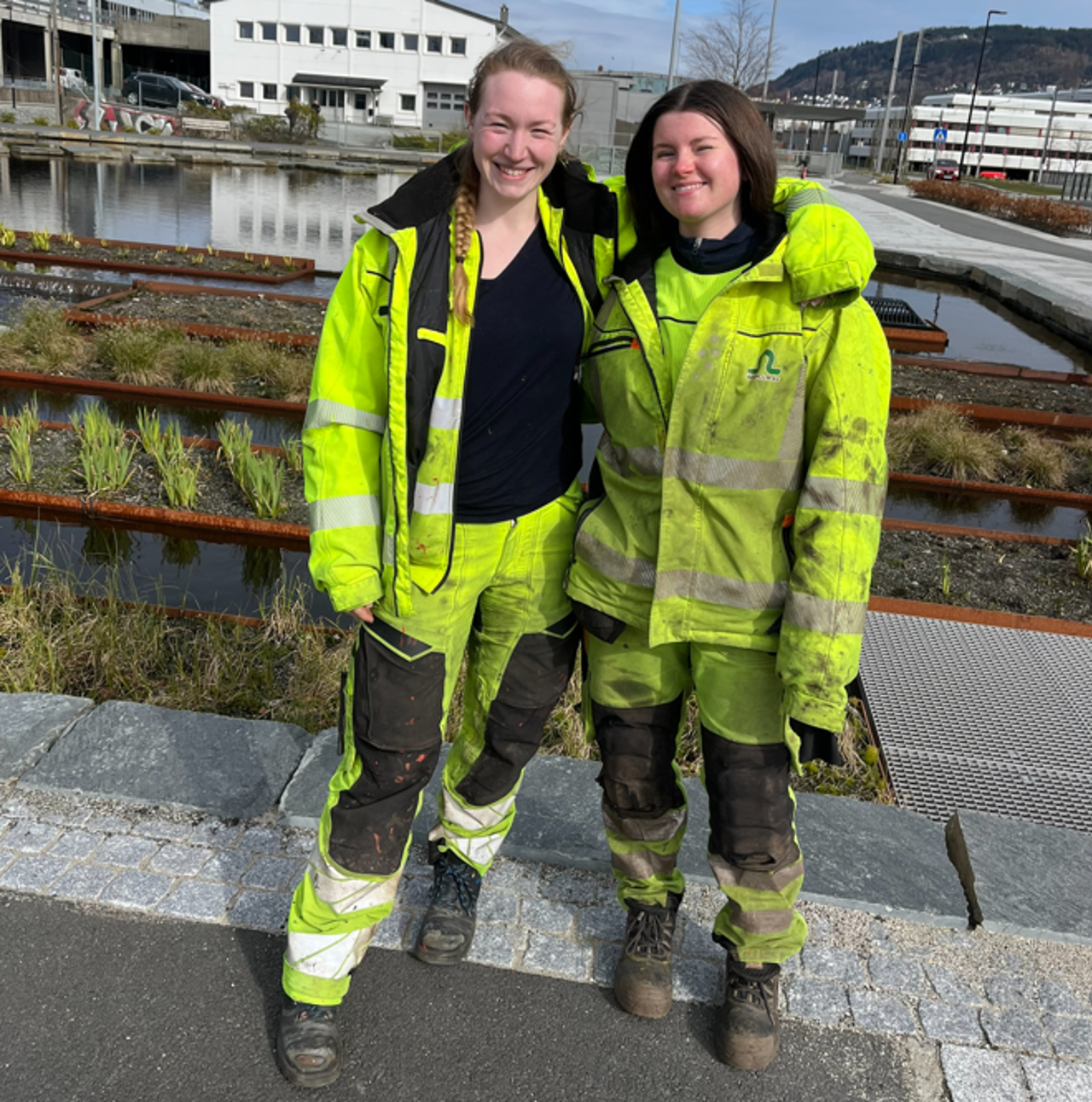 Biletet syner to kvinner - Emily Gabrielsen og Mariell Gaustad Hindenes - i gult synlegheitstøy med bassenget og kanalen på Mindemyren i bakgrunnen.